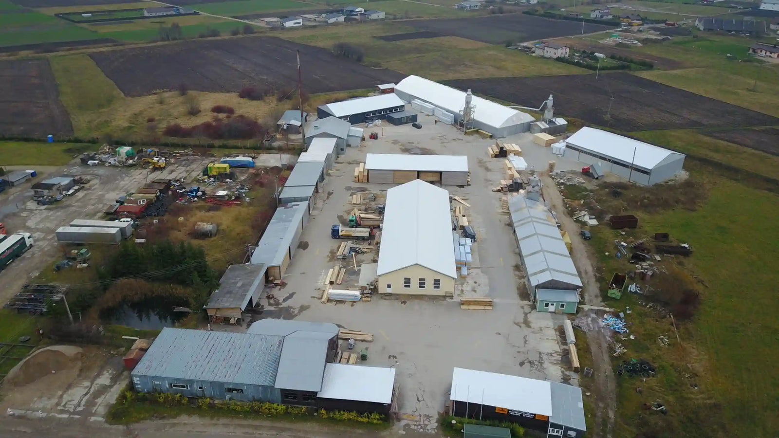 Imagen aérea de un complejo industrial rural. La fotografía muestra varios edificios grandes con techos de metal, algunos de ellos almacenes y otros talleres, rodeados de terrenos agrícolas. Hay vehículos y materiales de construcción dispersos alrededor de los edificios, indicando actividad industrial o de construcción.